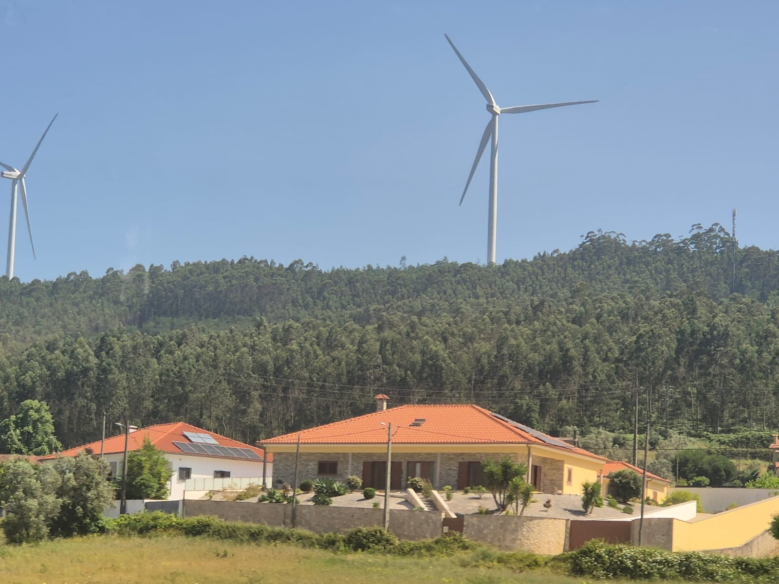 טורבינות רוח ענקיות ניצבות על רכס מיוער בצפון פורטוגל, מאחורי בתי מגורים עם גגות רעפים אדומים ופאנלים סולאריים. התמונה ממחישה את השילוב שבין מגורים כפריים לאנרגיה ירוקה באזור הררי וירוק.