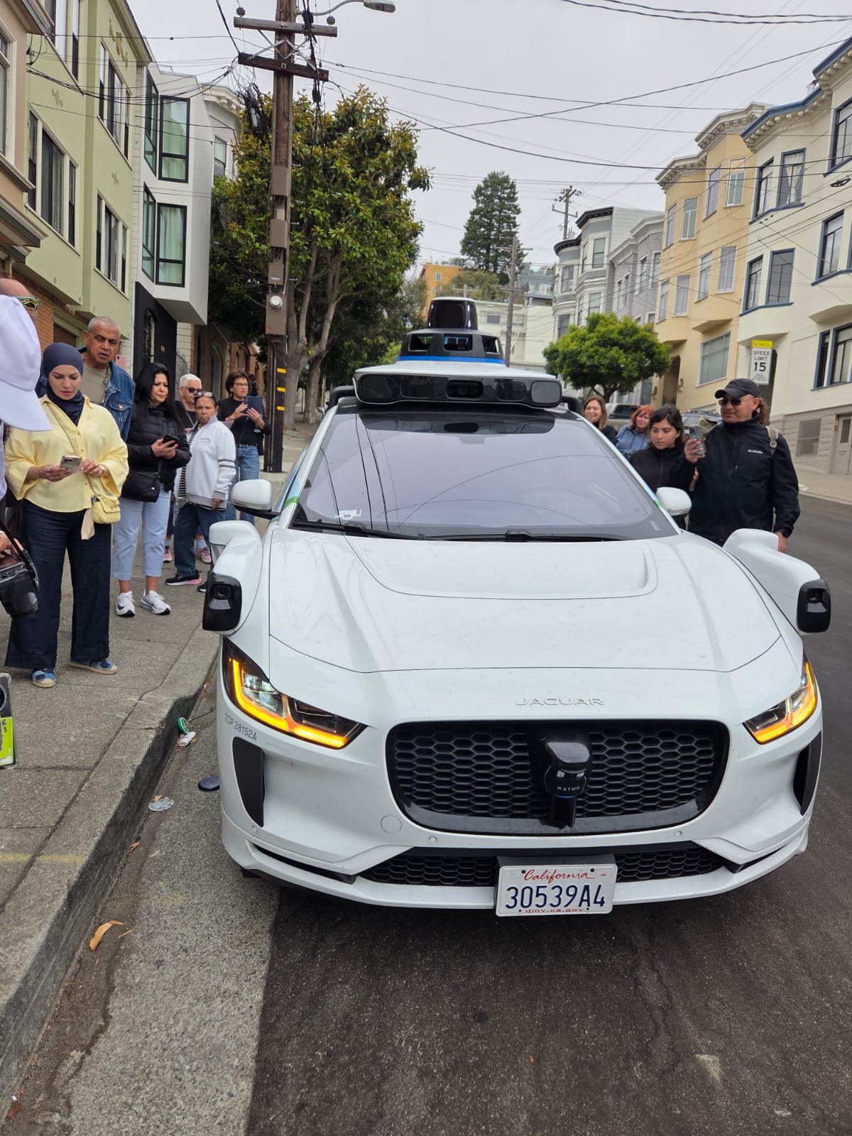 מונית אוטונומית לבנה מדגם יגואר ברחוב בסן פרנסיסקו, מוקפת בקהל סקרן