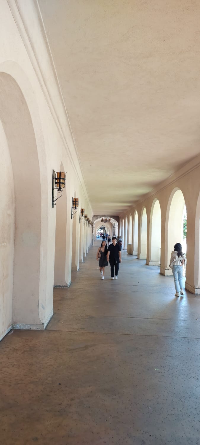 מבקרים פוסעים במסדרון מקושת ומוצל בסגנון ספרדי בפארק בלבואה, סן דיאגו