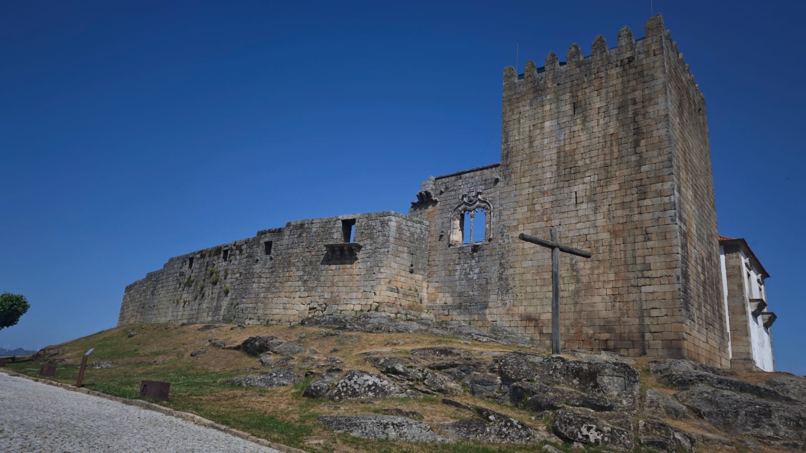 מצודת בלמונטה העתיקה – טיול לפורטוגל בעקבות היהדות. חומות אבן מרשימות עם מגדל שמירה ריבועי ומצלב עץ פשוט במרכז התמונה. השמיים הבהירים ברקע מדגישים את עוצמת המבנה ההיסטורי והקשר למורשת הדתית.