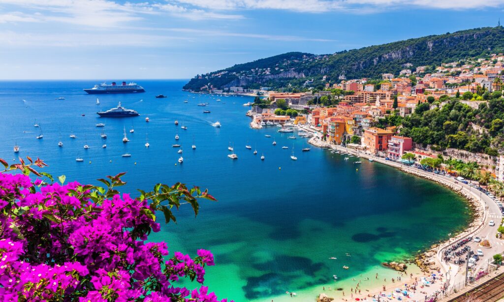 טיול מאורגן לריביירה האיטלקית והצרפתית – נוף חוף התכלת עם ים כחול עמוק, סירות ויאכטות, עיירת חוף צבעונית ופריחה סגולה בקדמת התמונה